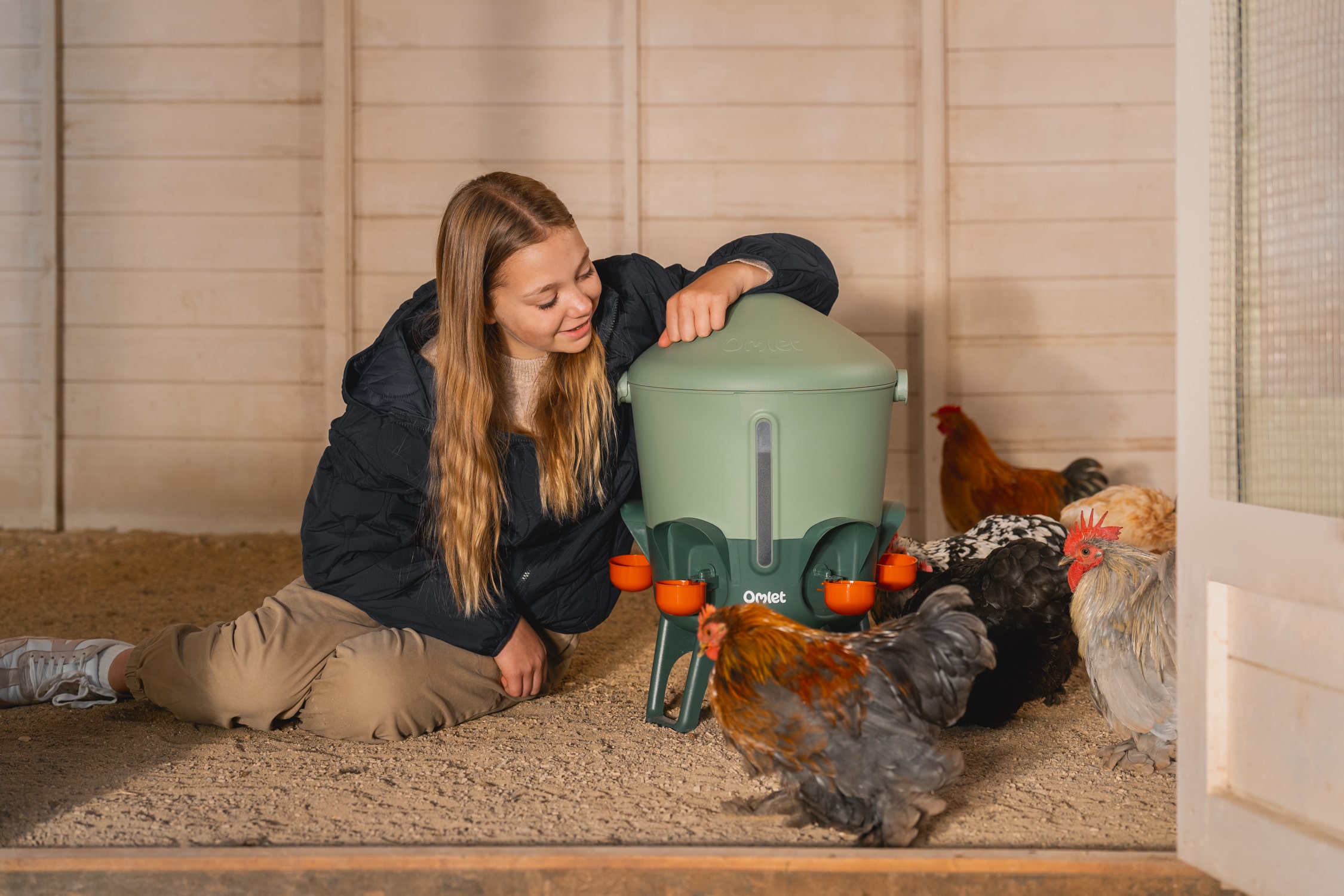girl sits next to a water trough with chickens drinking from cups