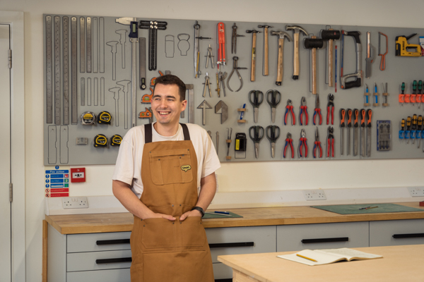 Ontwerpmanager Josh, staande in de werkplaats met gereedschap achter hem aan de muur, gekleed in een Omlet-werkschort.