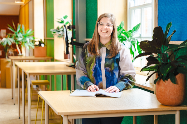 Rebecca, productontwerponderzoeker, stond bij een houten tafel met een notitieboekje en planten, glimlachend naar de camera.