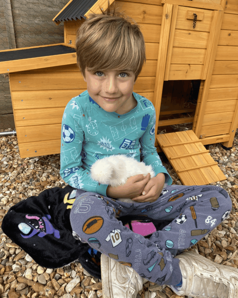 een jongetje met een kip op schoot zit voor het kippenhok